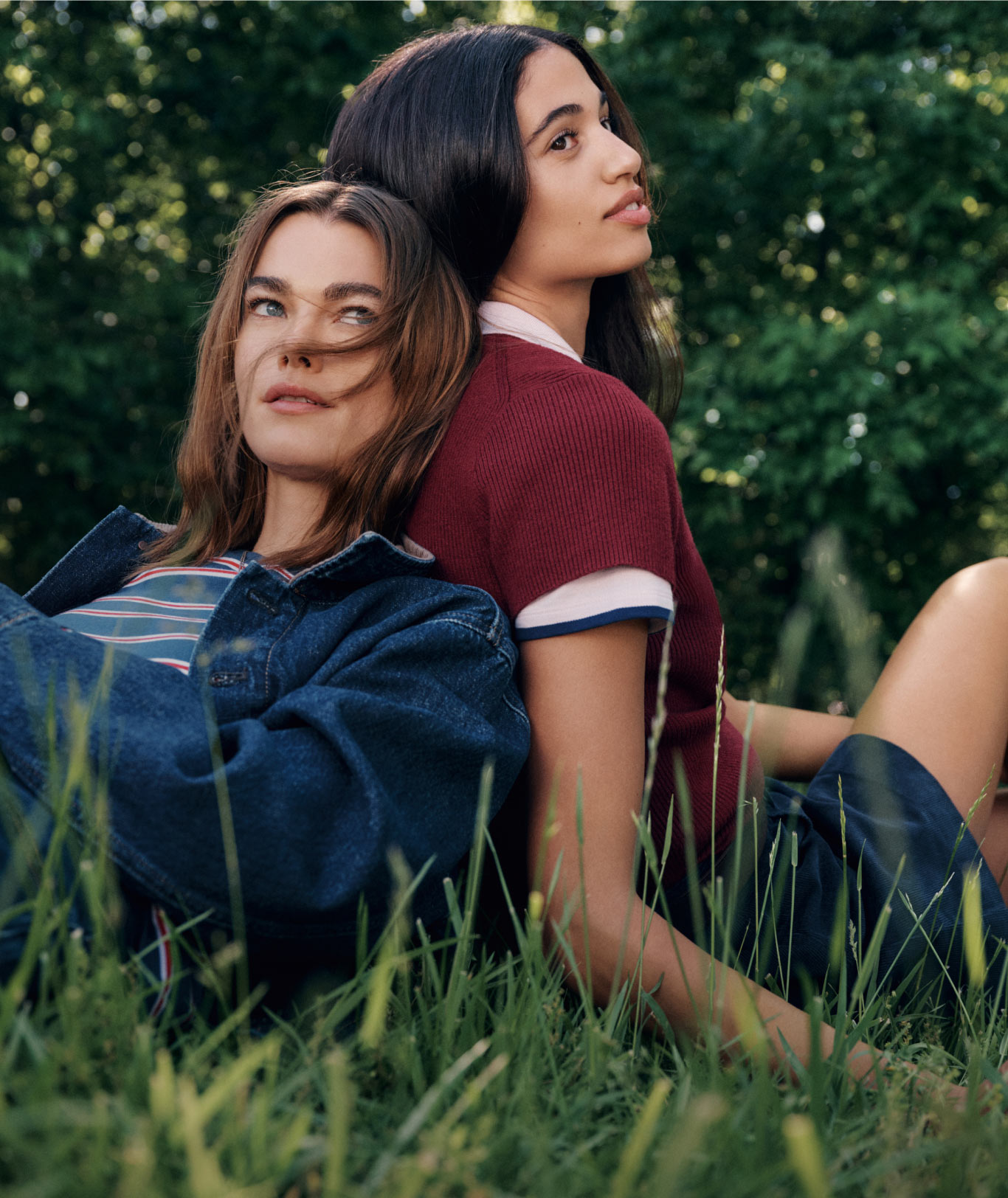 Two models in burgundy Tommy Hilfiger T-shirts and denim, sitting on the grass in a wooded outdoor environment.