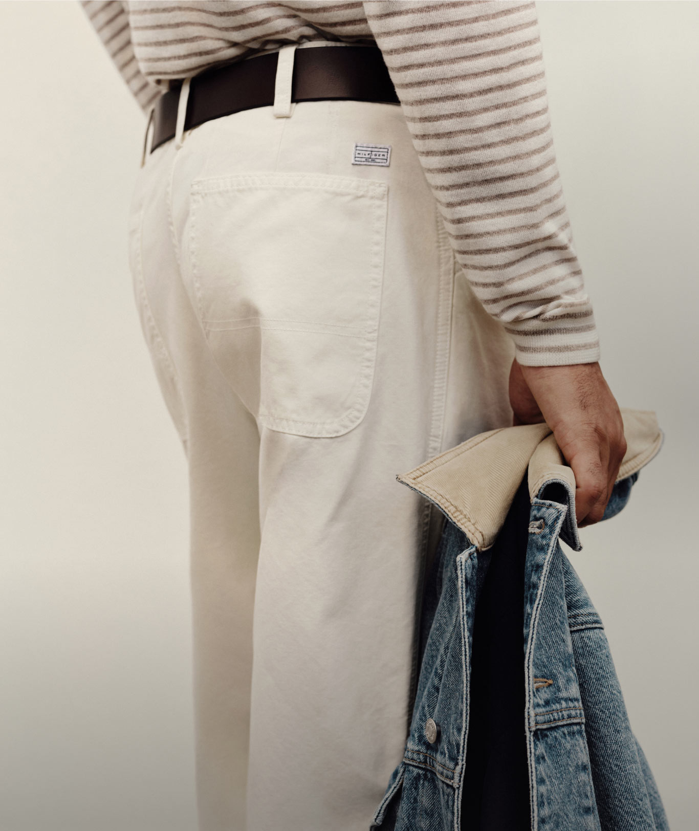 Close-up of a model wearing off-white pants and a striped Tommy Hilfiger top, holding a denim jacket.