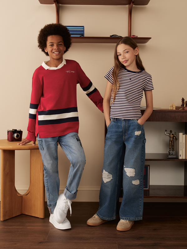 Two kids posing indoors, one wearing a red striped Tommy Hilfiger sweater with jeans and the other in a striped Tommy Hilfiger T-shirt with wide-leg ripped jeans.