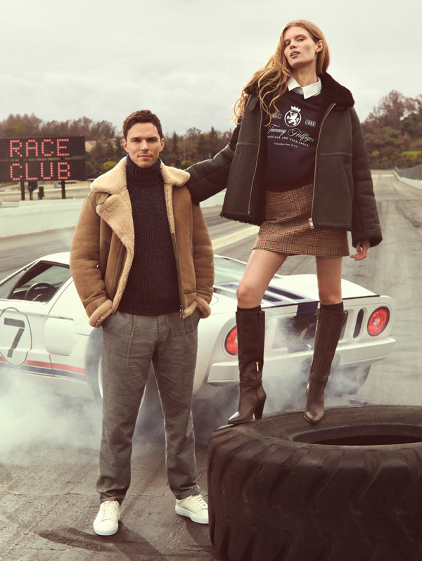 Man and woman posing on a racetrack in Tommy Hilfiger outerwear, with a sports car and large tire in the background.