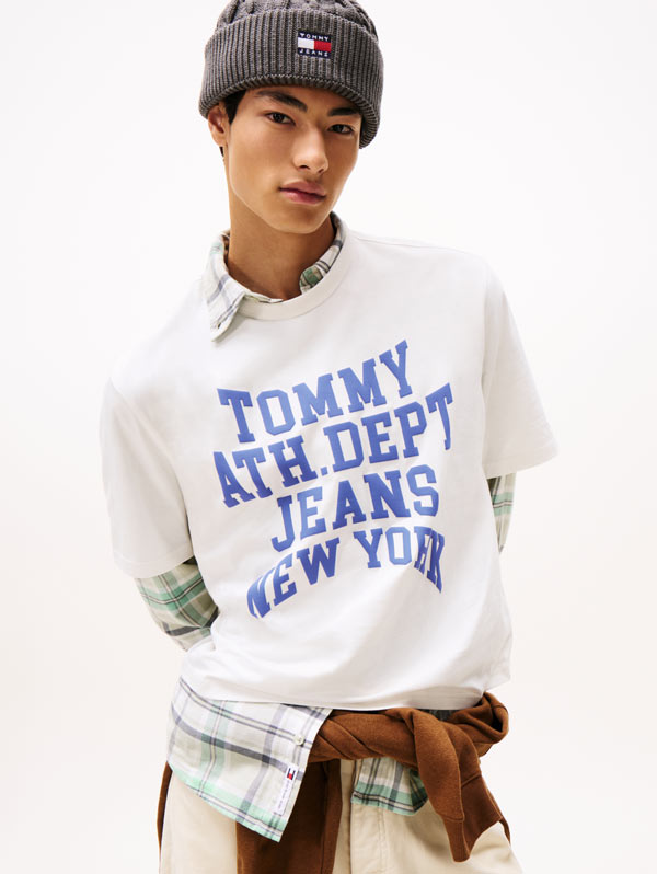 Man wearing a gray beanie and white Tommy Hilfiger graphic T-shirt with blue lettering, layered over a plaid shirt tied at the waist.