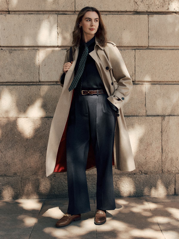 Woman standing outdoors wearing a long beige Tommy Hilfiger trench coat over wide-leg jeans and a dark top.