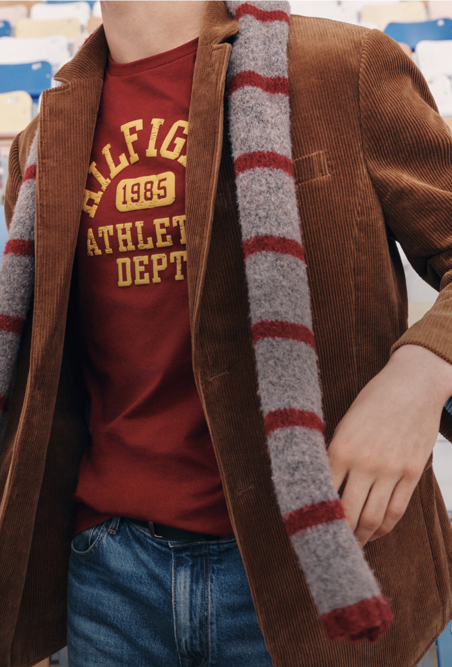 Model wearing a Tommy Hilfiger graphic tee, striped scarf, and brown jacket with denim, captured in a close-up outdoor setting.