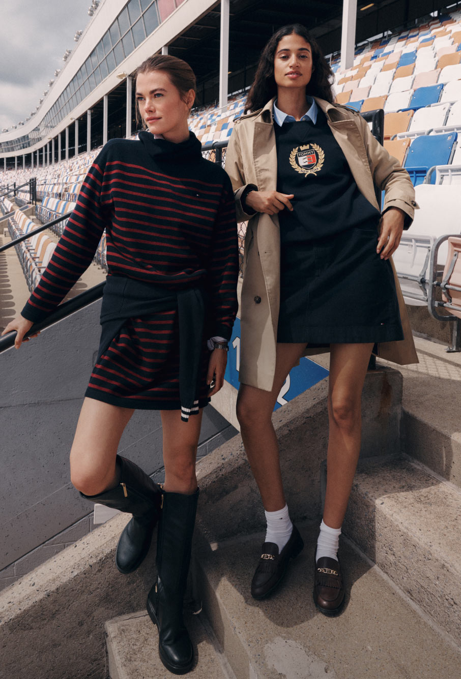 Two models wearing Tommy Hilfiger dresses, including a striped knit and a belted navy look, walking through a racetrack stadium.