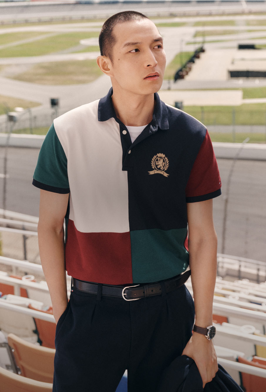 Male model in a colorblocked Tommy Hilfiger polo shirt with crest logo, standing by stadium seating with a confident pose.