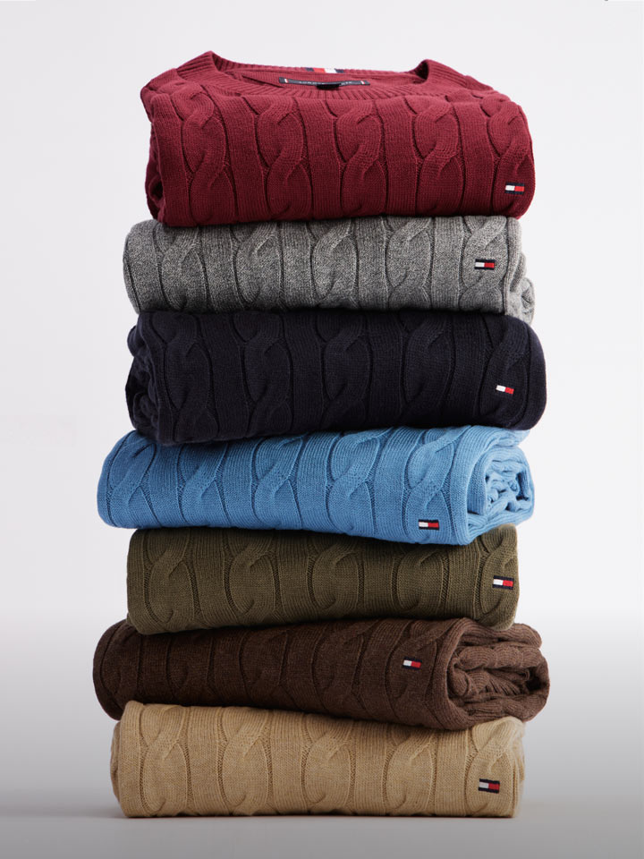 Stack of folded Tommy Hilfiger cable knit sweaters in assorted colors including red, gray, blue, brown, and beige.