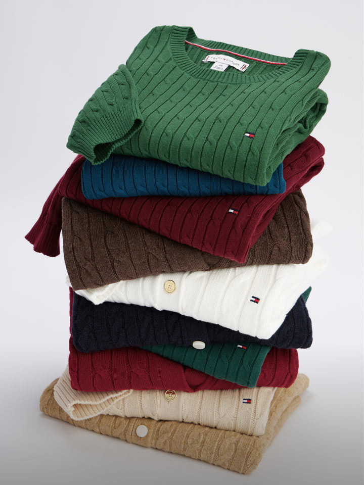 Stack of folded Tommy Hilfiger cable knit sweaters in assorted colors including green, red, white, navy, and beige.