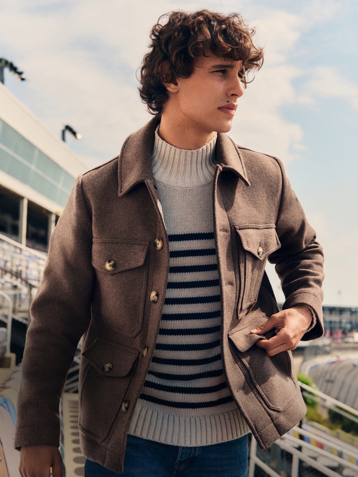 Man walking through stadium seating wearing a brown Tommy Hilfiger jacket over a striped sweater and jeans.