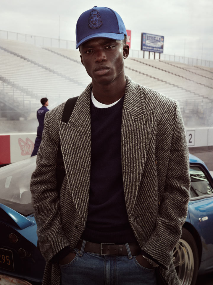 Man at a racetrack wearing a textured gray Tommy Hilfiger coat over a dark sweater and jeans, with a blue cap.