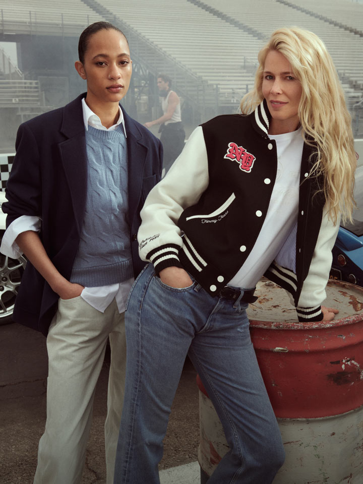 Two women at a racetrack, one in a navy blazer and white shirt, the other in a black and white Tommy Hilfiger varsity jacket with red accents.