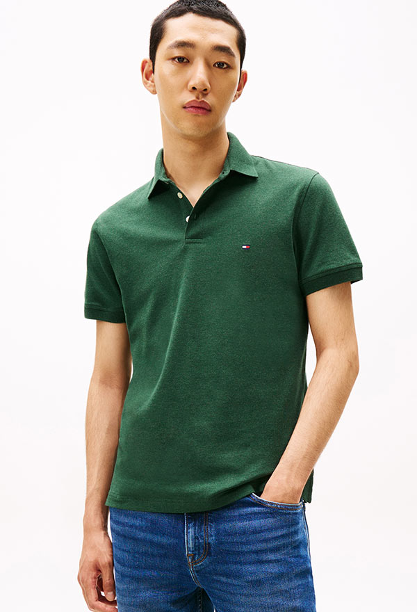 Model wearing a Tommy Hilfiger green short-sleeve polo shirt with subtle texture, styled with blue jeans, against a white background.