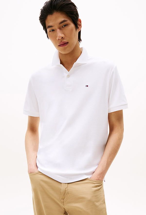 Model wearing a Tommy Hilfiger white short-sleeve polo shirt with embroidered logo, paired with khaki pants, posed against a white background.