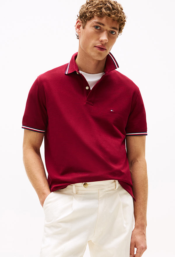 Model wearing a Tommy Hilfiger red short-sleeve polo shirt with white tipping, paired with cream pants, standing against a white background.