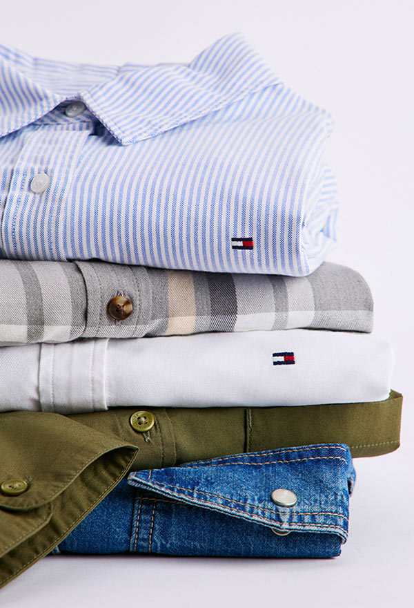 Stack of folded Tommy Hilfiger women's shirts in various styles and fabrics, including white, light blue, olive, and denim, showcasing collars and button details.