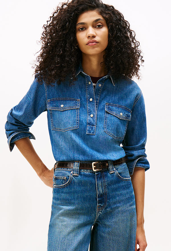 Model wearing a Tommy Hilfiger light indigo denim casual shirt with rolled sleeves, styled with blue jeans, standing against a white background.
