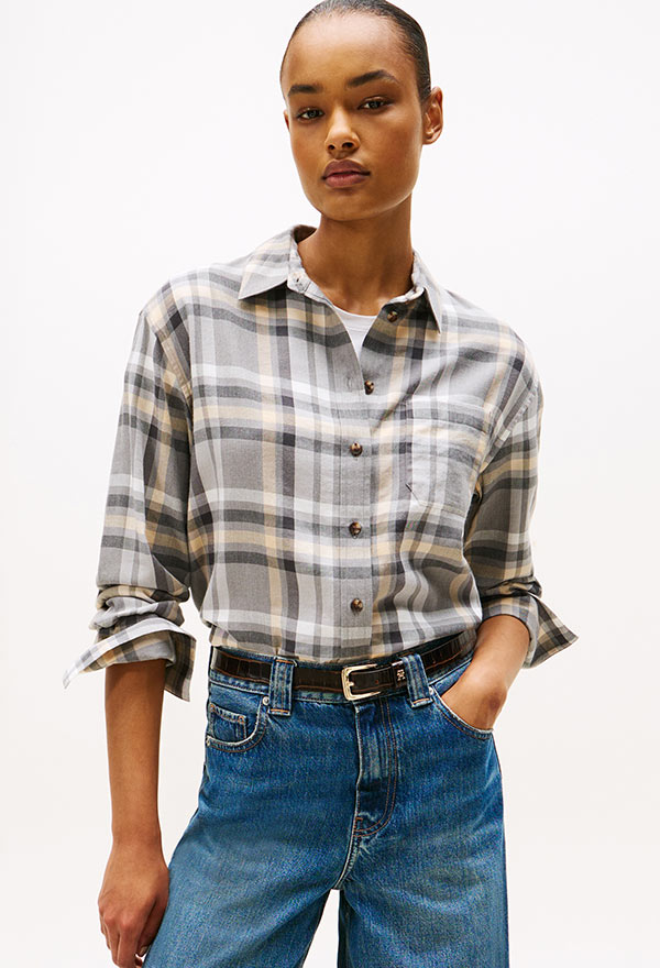 Model wearing a Tommy Hilfiger neutral plaid flannel shirt with rolled sleeves, styled with medium wash jeans, posed against a white background.
