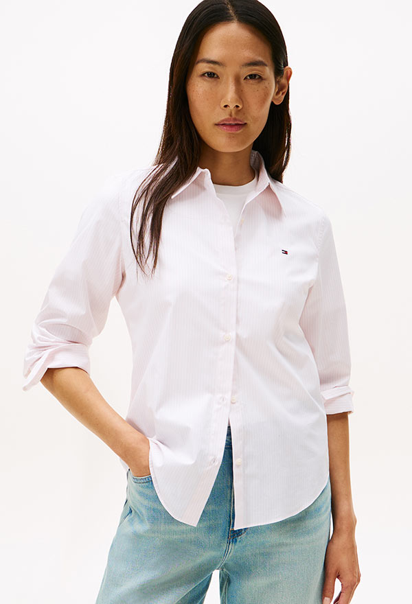 Model wearing a Tommy Hilfiger white oxford button-down shirt with rolled sleeves and light-wash jeans, standing against a white background.
