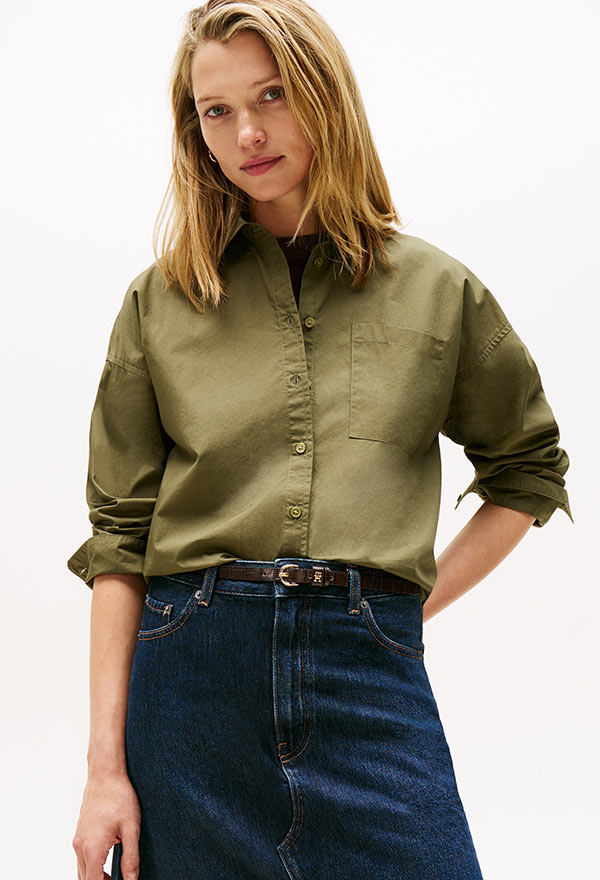 Model wearing a Tommy Hilfiger olive green poplin button-down shirt tucked into a dark denim skirt, posed against a white background.