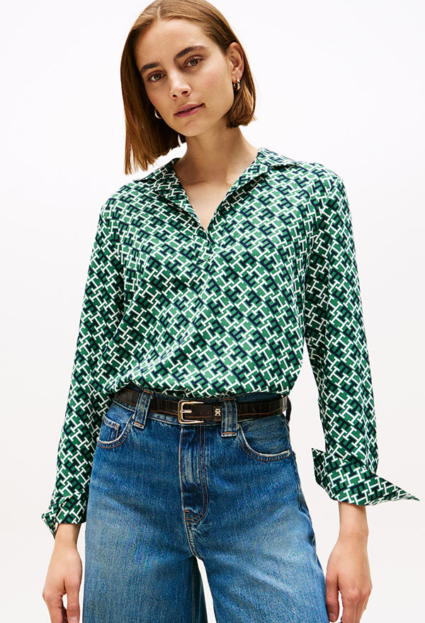 Model wearing a Tommy Hilfiger green and white printed button-down shirt with a geometric pattern, paired with blue jeans, standing against a white background.