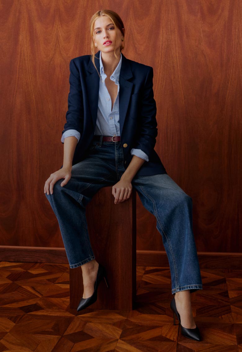 Woman wearing a navy blazer over a white shirt with barrel leg Tommy Hilfiger jeans, sitting against a wooden backdrop