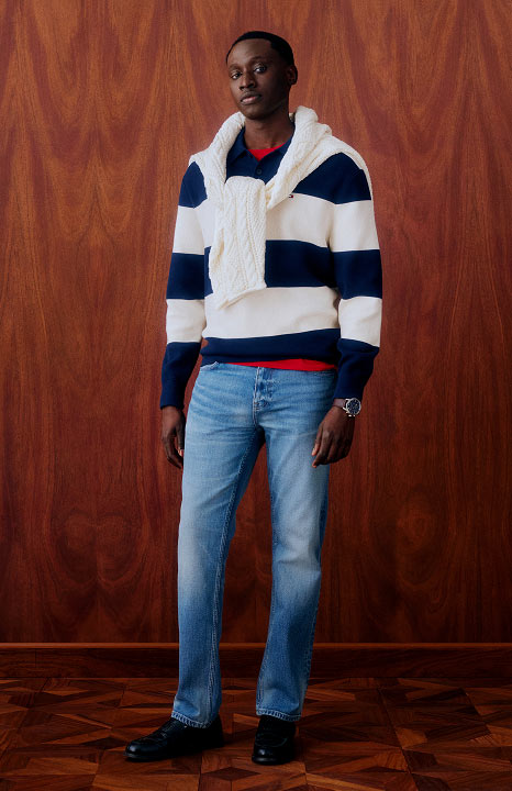 Man wearing a navy and white striped Tommy Hilfiger cable knit sweater over a red shirt with slim fit jeans, posing against a wooden backdrop.