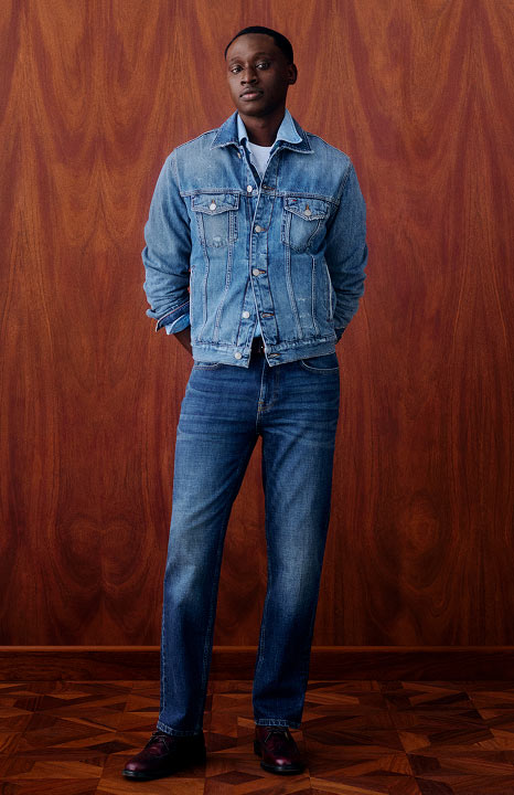Man wearing a light wash denim Tommy Hilfiger jacket over a chambray shirt with straight leg jeans, posing against a wooden backdrop.