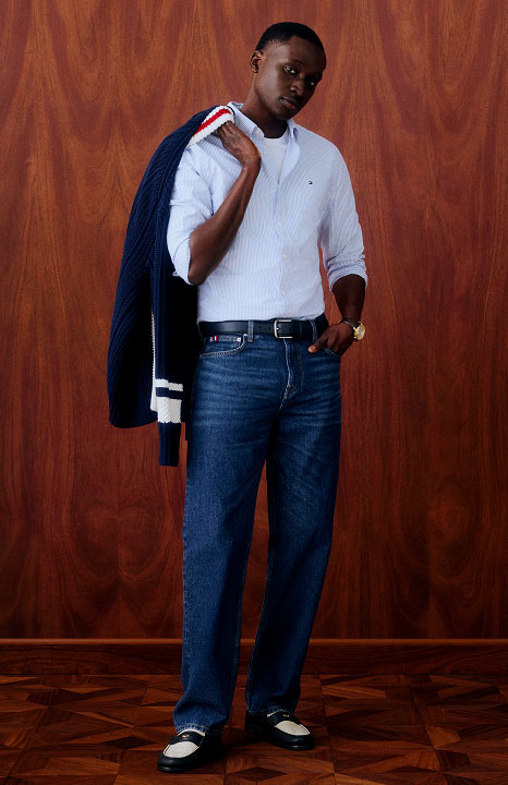 Man wearing a white shirt with a black and white Tommy Hilfiger varsity jacket draped over his shoulder, paired with relaxed fit jeans, posing against a wooden backdrop.