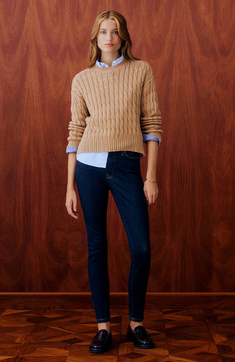 Woman wearing a tan cable knit Tommy Hilfiger sweater over a white shirt with skinny fit jeans, posing against a wooden backdrop.