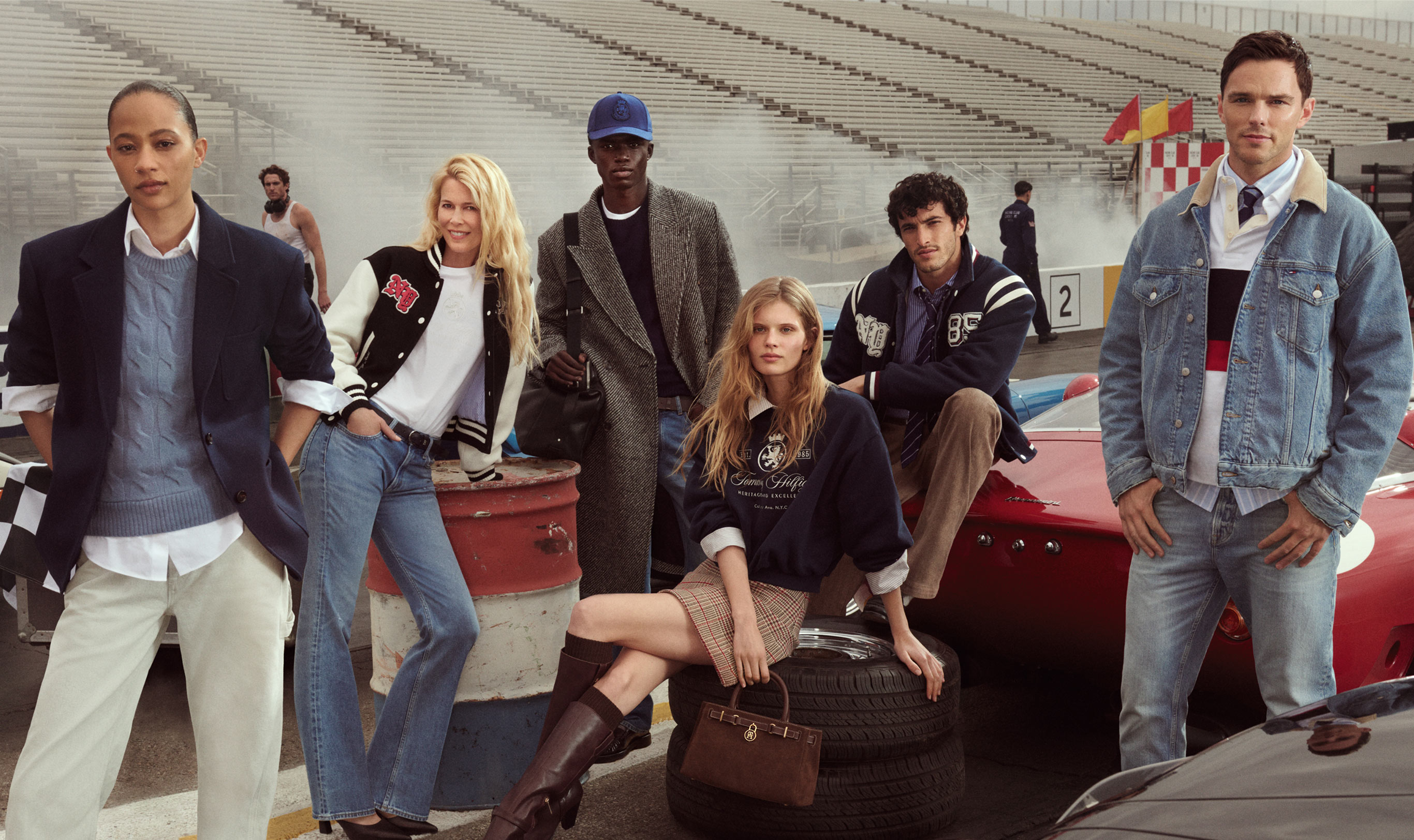 A group of men and women in front of race cars, wearing Tommy Hilfiger.