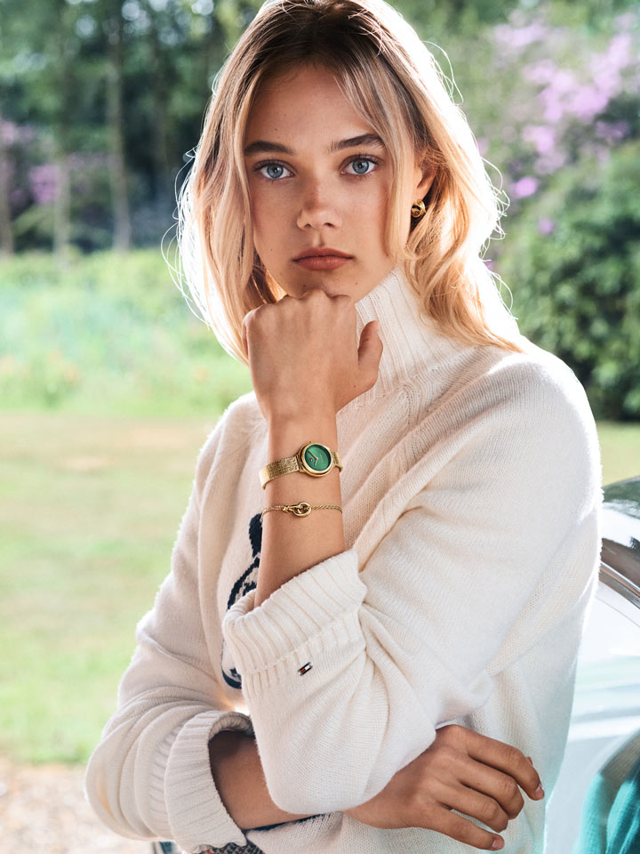 Model wearing a light blazer and gold-tone Tommy Hilfiger jewelry, posing with hand near face in a softly lit environment.