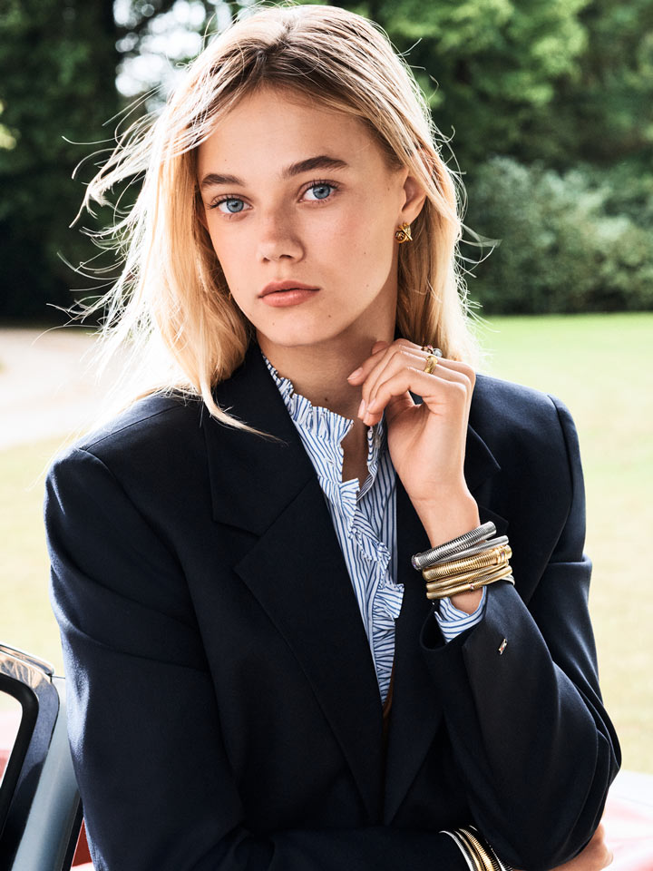 Model in a navy blazer styled with layered Tommy Hilfiger bracelets and rings, seated beside a car outdoors.