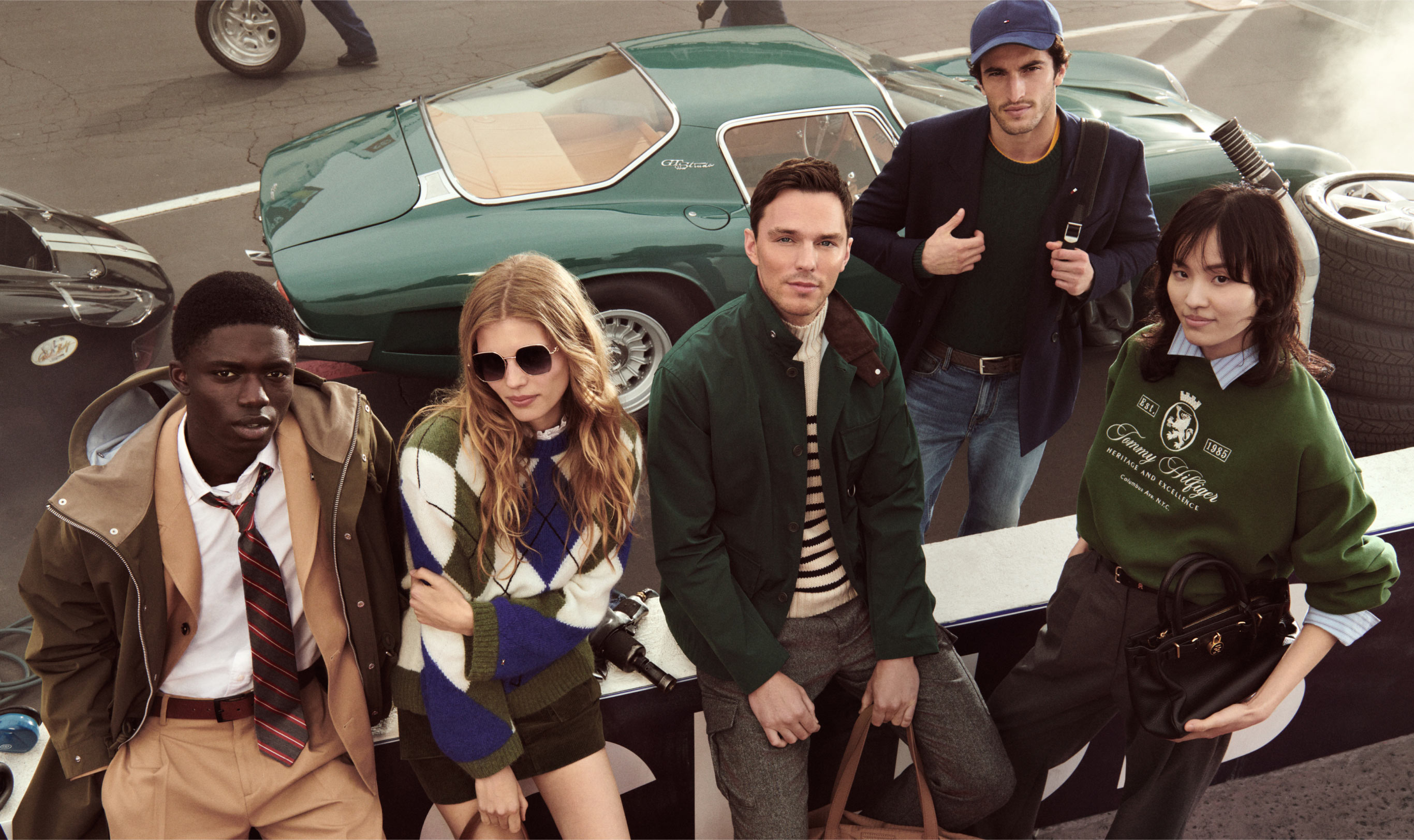 Group of models wearing Tommy Hilfiger global-inspired styles, including varsity jackets, sweaters, and polos, standing in front of vintage cars.