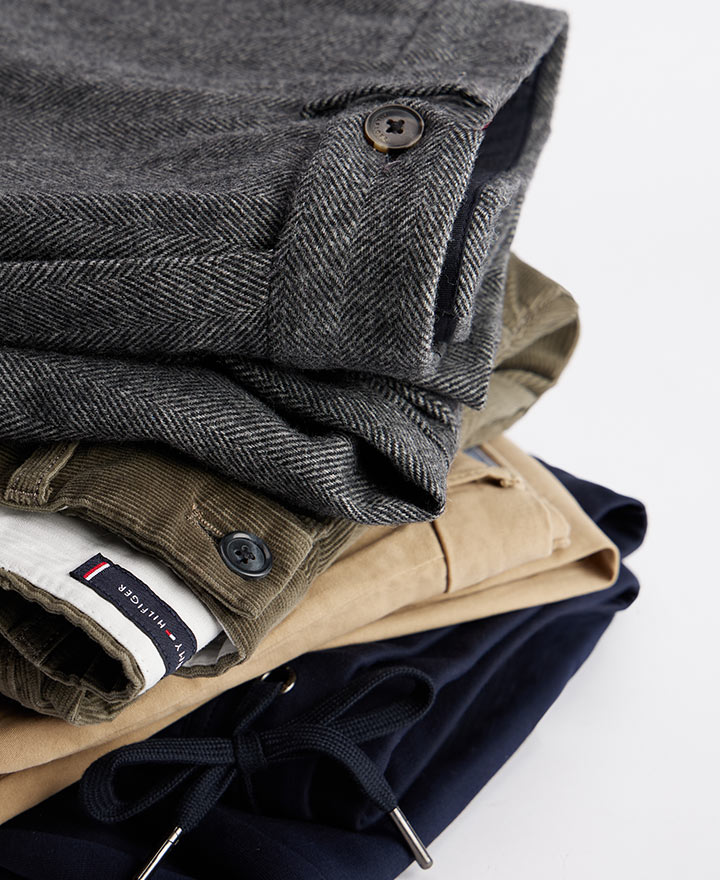 Stack of folded Tommy Hilfiger men’s pants and trousers in various shades including grey, tan, and navy, highlighting logo waistbands and belt loops.
