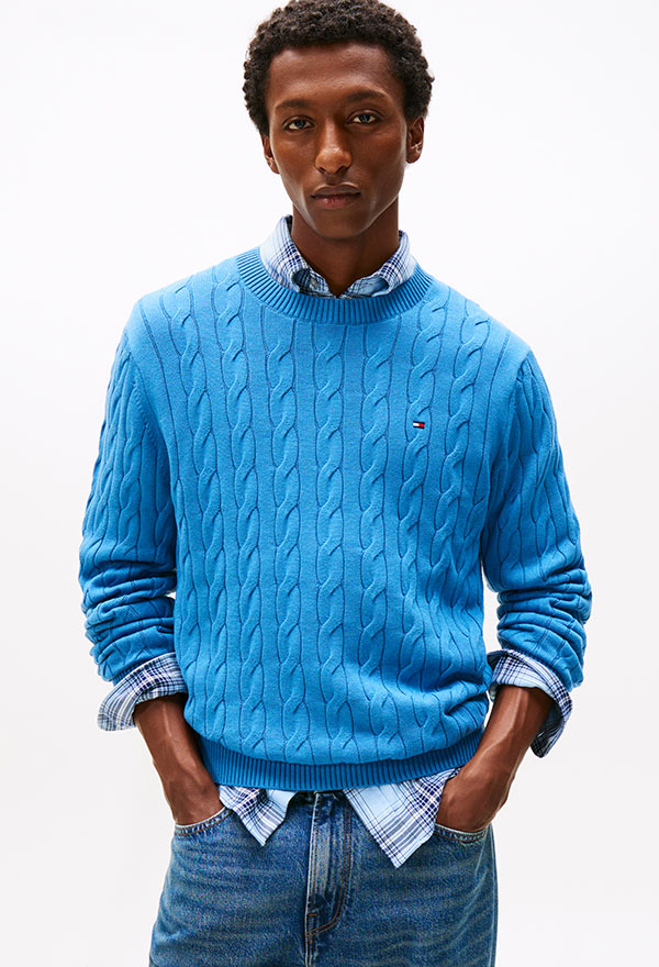 Model wearing a Tommy Hilfiger light blue cable-knit crewneck sweater with dark jeans, standing against a white background.