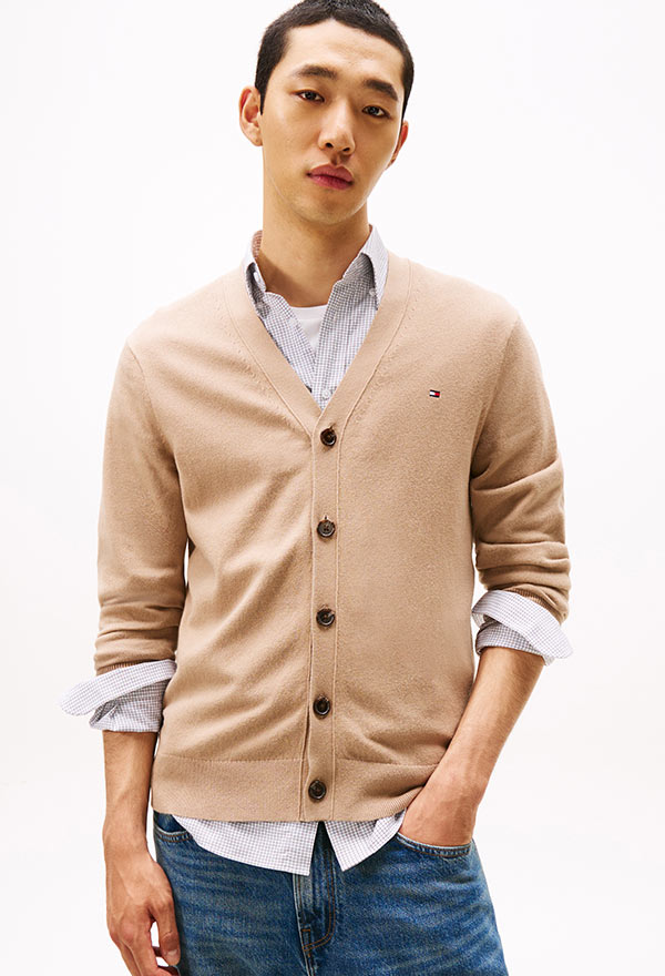 Model wearing a Tommy Hilfiger beige button-up cardigan layered over a white T-shirt, styled with blue jeans, posed against a white background.