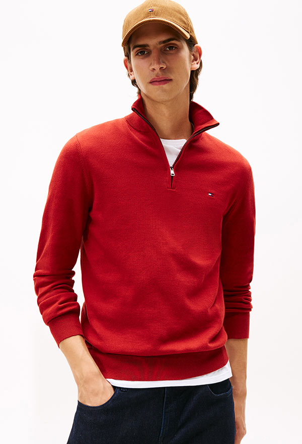Model wearing a Tommy Hilfiger red quarter-zip pullover sweater with contrast collar trim and dark pants, posed against a white background.