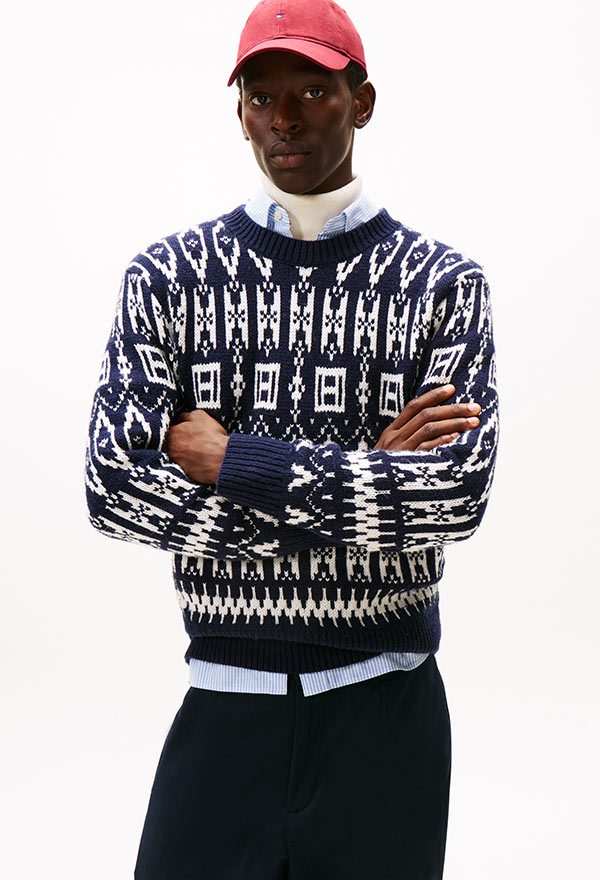 Model wearing a Tommy Hilfiger navy and cream fair isle patterned crewneck sweater with a red baseball cap and dark jeans, posed against a white background.