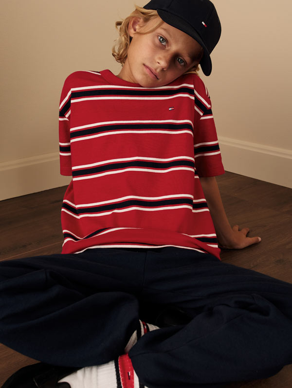 Model seated indoors in a red and white striped Tommy Hilfiger knit top and navy pants, wearing a baseball cap tilted to the side.