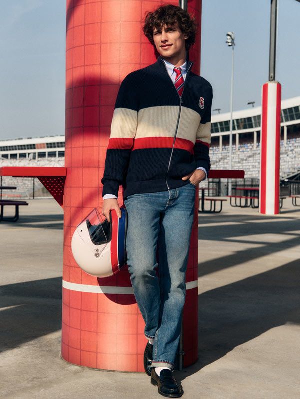 Model leaning against a red column in a colorblock striped Tommy Hilfiger sweater and jeans, holding a racing-style helmet.