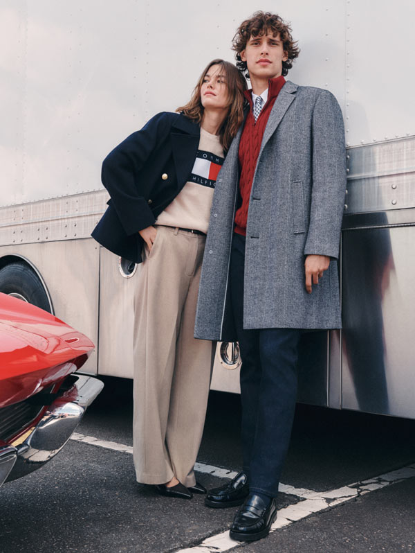 Two models wearing Tommy Hilfiger preppy styles—one in a diamond-patterned sweater and pleated skirt, the other in a long wool coat with a red turtleneck—standing near a vintage race car backdrop.