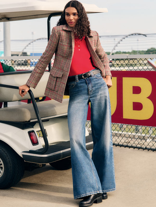 Model standing beside a car wearing a cropped red top, plaid jacket, and wide-leg Tommy Hilfiger jeans in a retro-inspired pit stop setting.