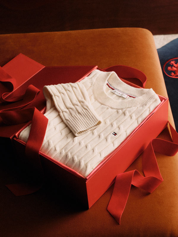 Folded cream cable-knit Tommy Hilfiger sweater and matching socks presented in a red gift box with ribbon, placed on a tan leather surface.