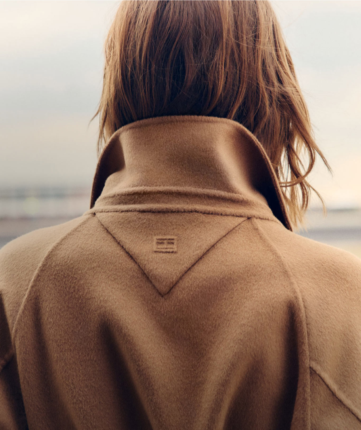 Models wearing Tommy Hilfiger outerwear one in a structured coat and trousers walking near a red convertible, the other seen from behind in a cozy camel coat with oversized collar.