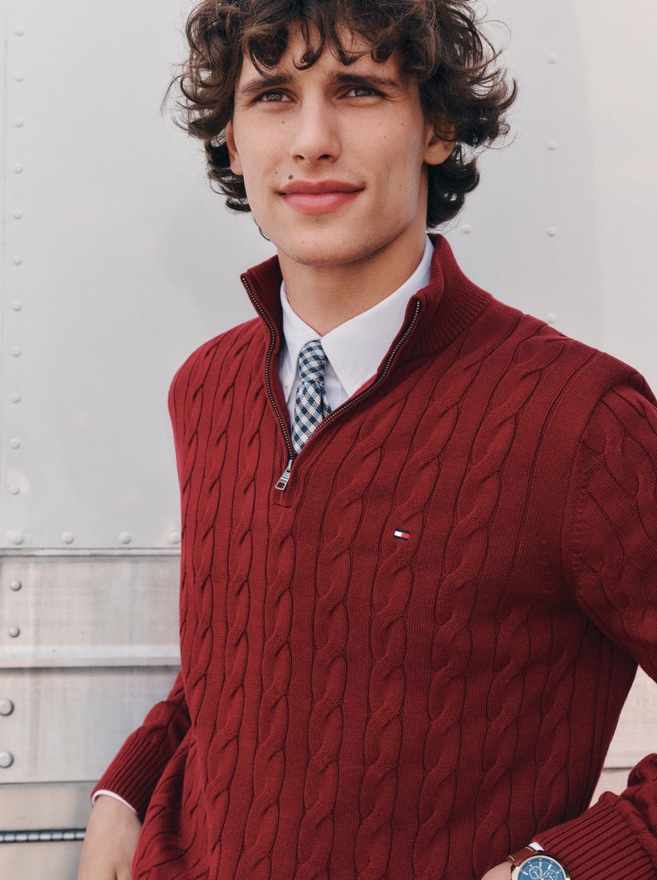 Model wearing a red Tommy Hilfiger cable-knit sweater over a white shirt and striped tie, shot waist-up against a light wall.