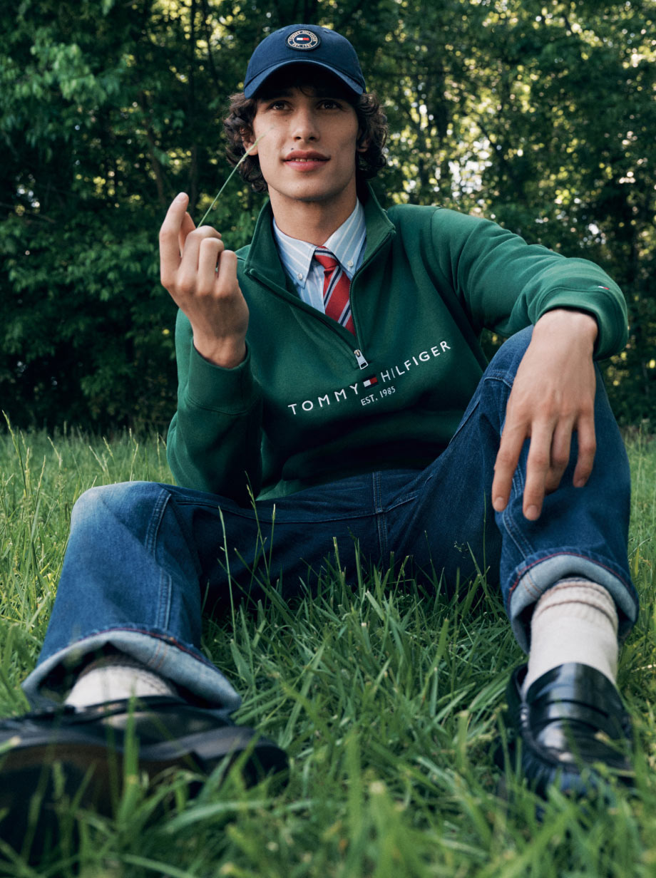 Model sitting on grass in a green Tommy Hilfiger sweater over a collared shirt and tie with jeans and a navy cap, outdoors in a park.