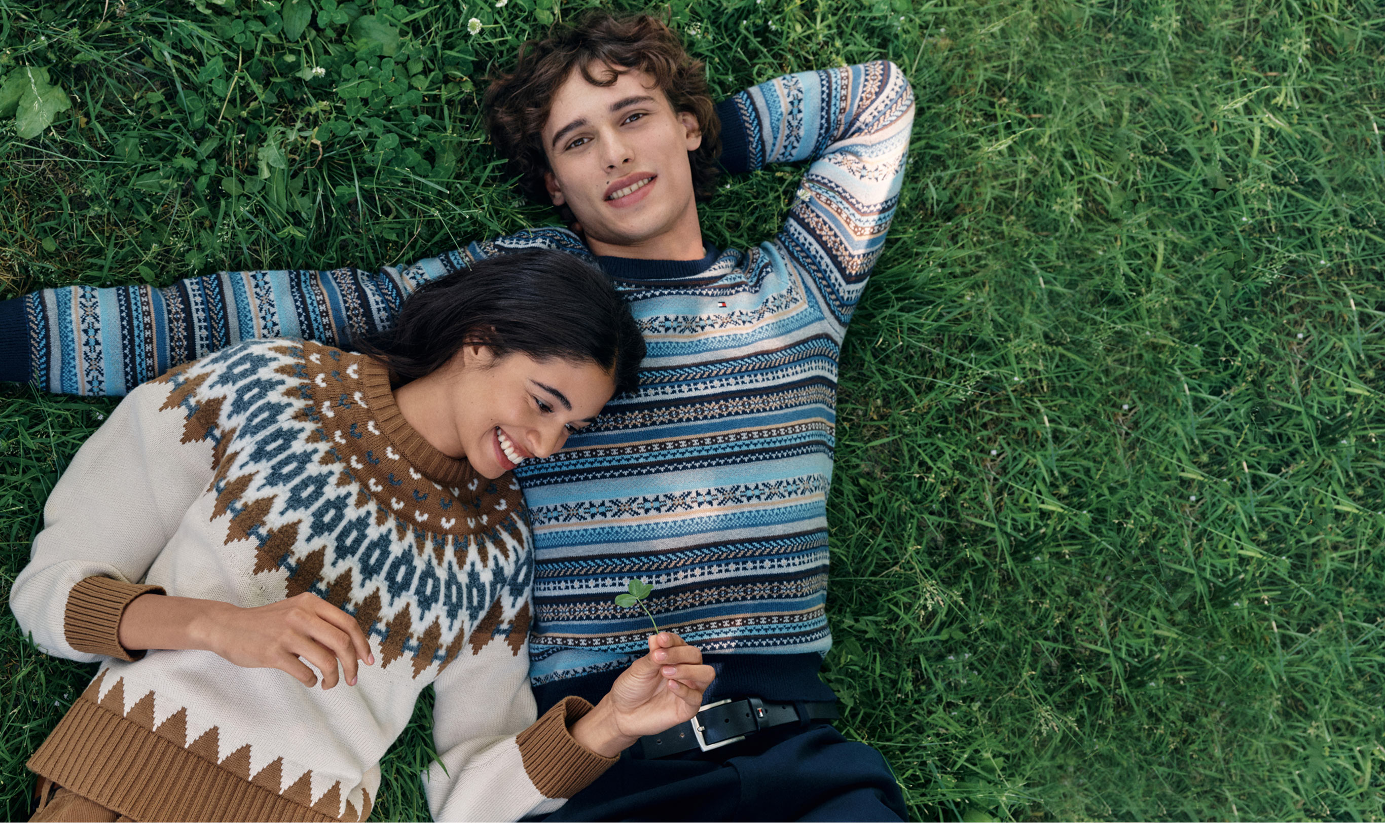 Two models lying on grass wearing Tommy Hilfiger knit sweaters: one in a cream fair isle and the other in a blue striped crewneck.