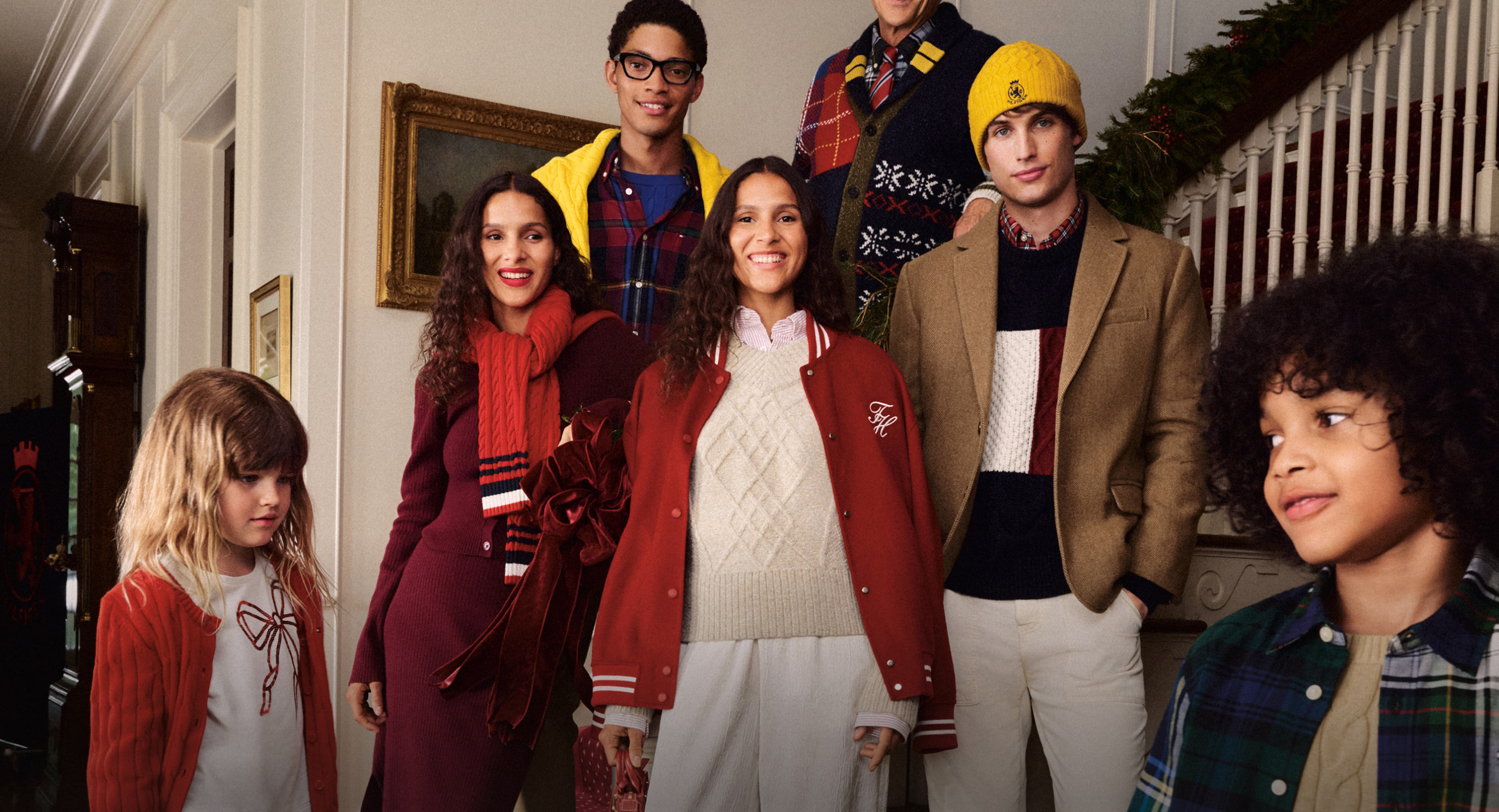 Group of models dressed in Tommy Hilfiger holiday outfits, including sweaters, scarves, blazers, and festive layers, gathered on a staircase in a warmly decorated home.