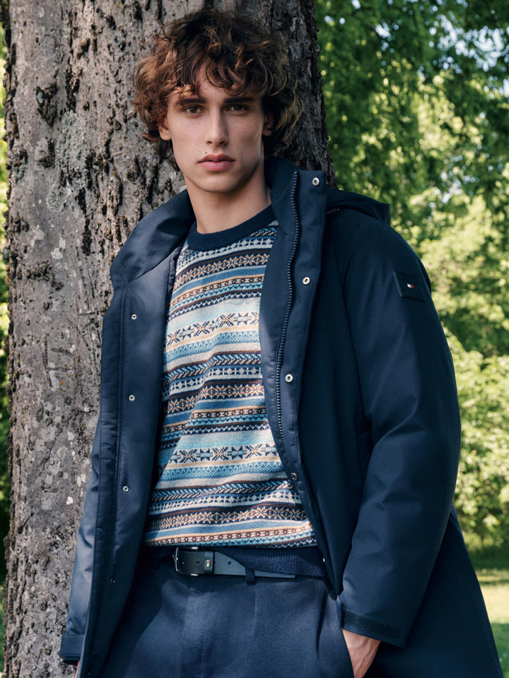 Model leaning against a tree in a navy Tommy Hilfiger parka over a multicolor fair isle sweater with relaxed navy trousers, outdoors in a park.