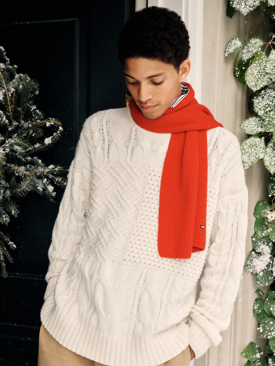 Model wearing a white Tommy Hilfiger cable-knit sweater with a bright red scarf, indoors beside holiday greenery.