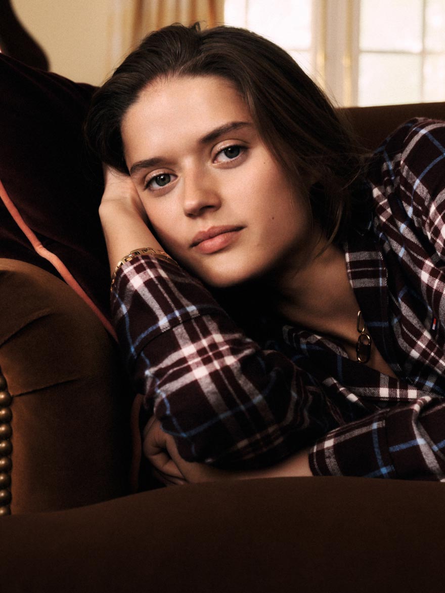 Close-up of a model lounging on a sofa in a navy-and-burgundy Tommy Hilfiger plaid flannel pajama shirt, indoors near a piano.